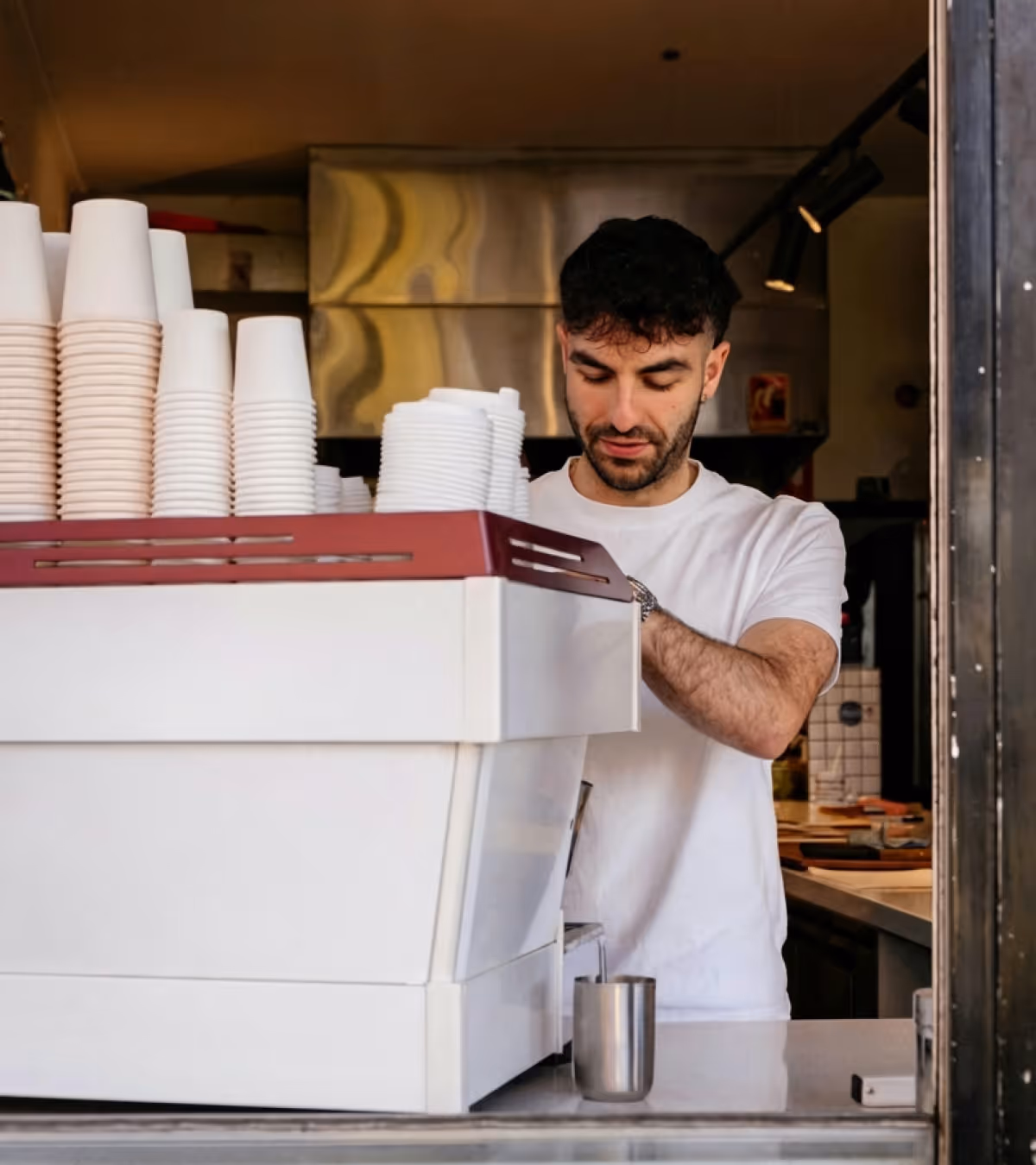 Barista preparing a coffee behind a counter with stacks of disposable cups next to an espresso machine.