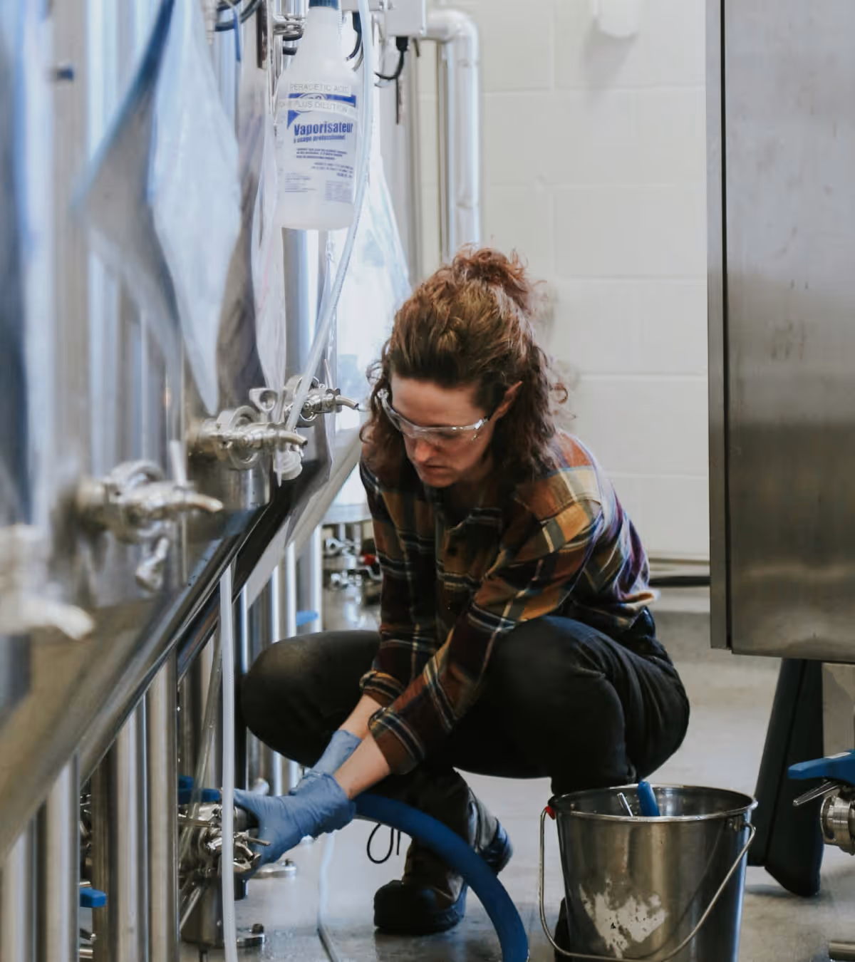 Person wearing safety glasses and gloves kneeling while working with a valve on a stainless steel brewing tank connected to a blue hose.
