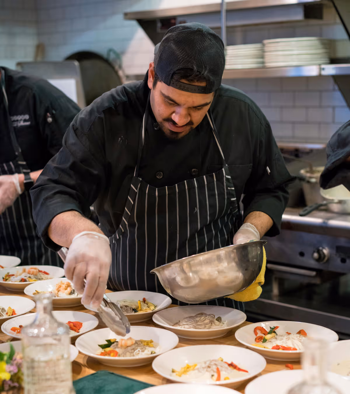 Chef wearing a black cap and striped apron carefully plating shrimp pasta in a professional kitchen.