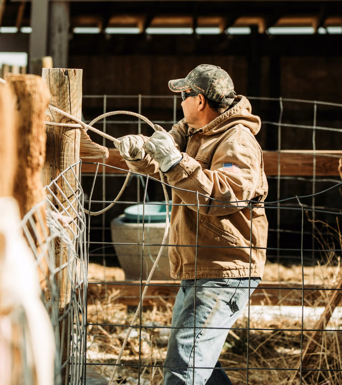 Man wearing gloves and a camo cap pulling a rope around a wooden post inside a fenced area on a farm.