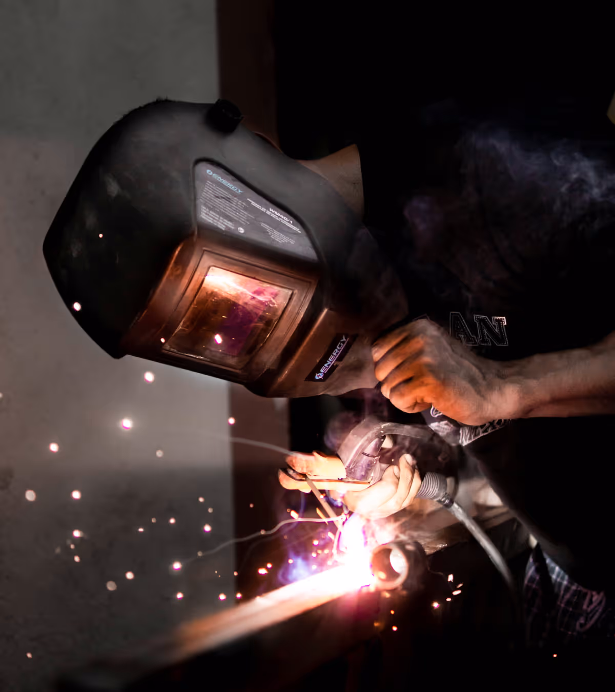 Welder wearing a protective helmet and gloves welding metal with sparks and smoke visible.