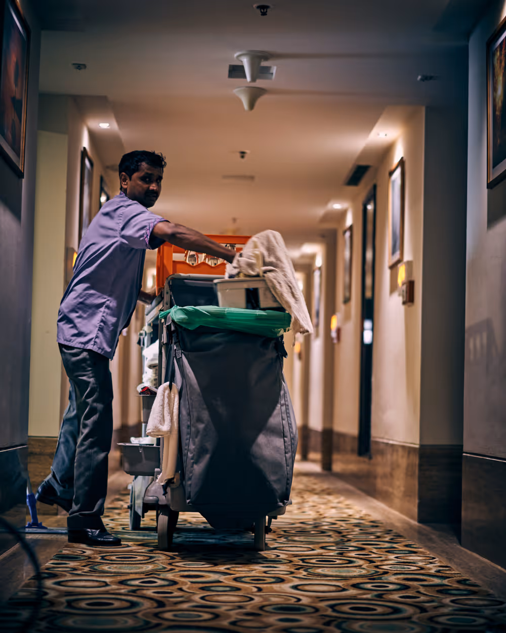 Hotel staff pushing a housekeeping cart loaded with cleaning supplies through a carpeted hallway.